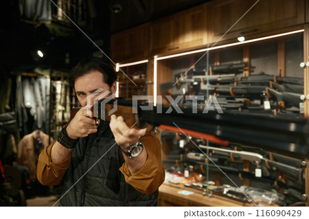 Weapon shop owner aiming with gun while standing over showcase gun rack 116090429