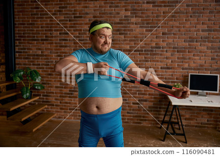 Comic chubby man in sportswear fooling around with fitness equipment Comic chubby man in sportswear fooling around with fitness equipment 116090481