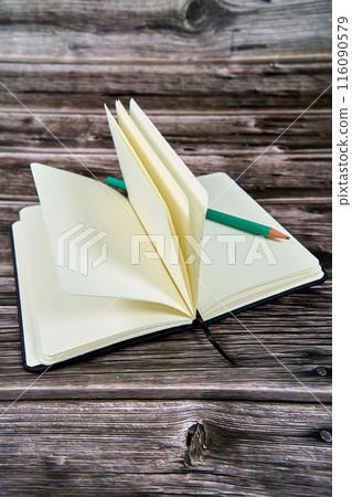 Open notebook with blank pages and a green pencil on a wooden table.  116090579