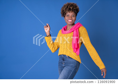 Young pretty black woman dancing with afro curly hair smiling in fuchsia scarf and yellow sweater on blue background with copy space. Happiness concept. Young pretty black woman dancing with afro curly hair smiling in fuchsia scarf and yellow sweater on blue background with copy space. Happiness concept. 116091900