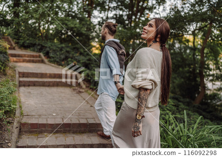 Outdoor shot of young couple in love walking on pathway through grass field. Young couple strolling in the park. Trendy couple with tattoos walking in a park. Botanic Garden. 116092298