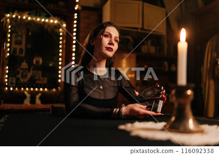 Mysterious wizard with black nails shuffling deck of tarot cards by light of burning candles. Mysterious clairvoyant performing esoteric rite, forecasting fate. Concept of esoteric, fortune telling. 116092338