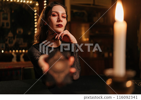Portrait of mysterious fortune teller asking client to move deck of cards for layout during session, in dark esoteric room by light of burning candle, looking at camera with serious expression. 116092341