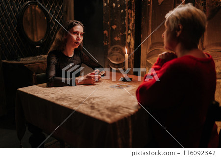 Rear view of fortuneteller doing Tarot card reading and talking to unhappy adult woman client about destiny in dark esoteric room by light of burning candle. Concept of prophecy, prediction. 116092342