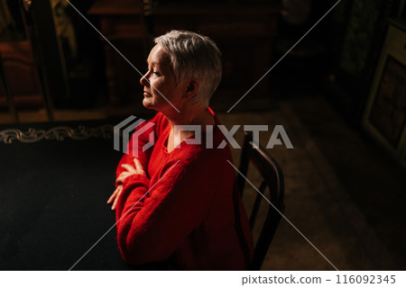 High-angle view of elegant gray-haired mature adult woman in casual red sweater sitting at table in dark room by light of burning candle, pensive looking away. High-angle view of elegant gray-haired mature adult woman in casual red sweater sitting at table in dark room by light of burning candle, pensive looking away. 116092345