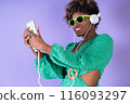 Young pretty black woman with curly hair wearing sunglasses and listening to music on her smartphone with headphones on a lilac background, with copy space. Happiness concept. 116093297