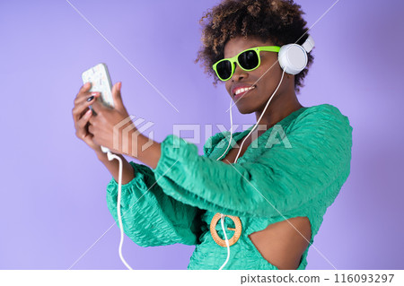 Young pretty black woman with curly hair wearing sunglasses and listening to music on her smartphone with headphones on a lilac background, with copy space. Happiness concept. Young pretty black woman with curly hair wearing sunglasses and listening to music on her smartphone with headphones on a lilac background, with copy space. Happiness concept. 116093297