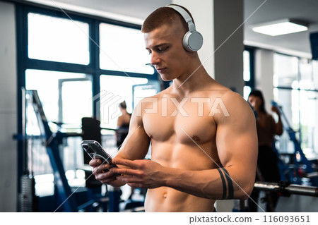 A shirtless man in a gym, wearing headphones, looks at his phone 116093651