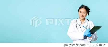 Smiling asian medical worker, female physician writing down patient info, holding pen and clipboard, standing in uniform over blue background 116094814