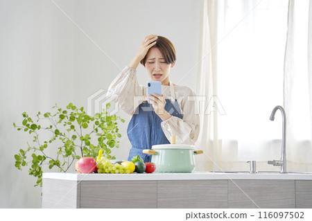 A woman looking at her smartphone while worrying in the kitchen A woman looking at her smartphone while worrying in the kitchen 116097502