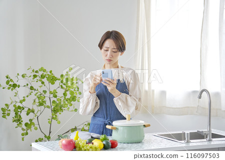 A woman operating a smartphone while worrying in the kitchen A woman operating a smartphone while worrying in the kitchen 116097503