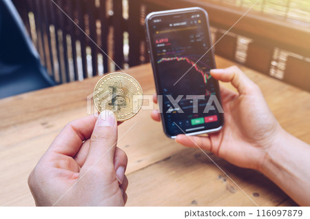 Someone checking Bitcoin price chart on digital exchange on smartphone while holding Bitcoin token. 116097879