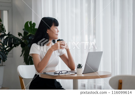 Young woman holding a coffee cup, sitting at a table with a laptop, glasses, and a small plant, looking out the window. 116099614