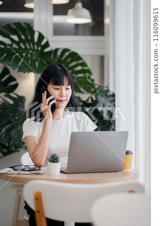 Young woman multitasking, working on a laptop and talking on the phone in a bright, modern home office with green plants. 116099615