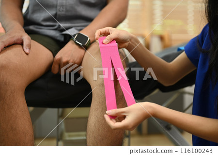 Cropped shot female physiotherapist applying pink kinesio tape on patient knee. Physical therapy and rehabilitation concept 116100243