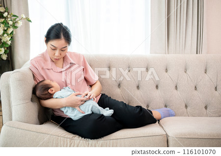 A young Asian mom comforting her newborn baby son to sleep on the couch together in the living room. 116101070