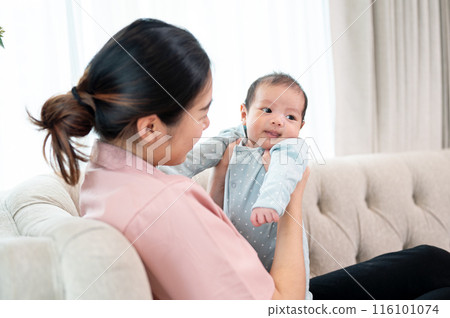 A happy Asian mom holding her adorable newborn baby son on a couch in the living room. 116101074