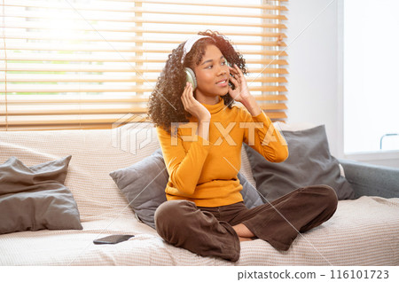 An attractive, cheerful Black woman is enjoying listening to music on headphones on the couch. 116101723