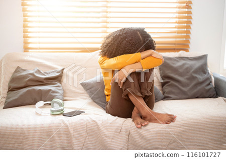 A depressed, upset young Black woman is sitting on the sofa, crying, feeling lonely and sad. 116101727