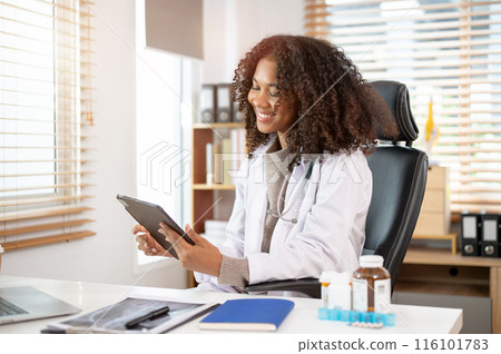 An attractive Black female doctor in a white gown is using her digital tablet in her office. An attractive Black female doctor in a white gown is using her digital tablet in her office. 116101783