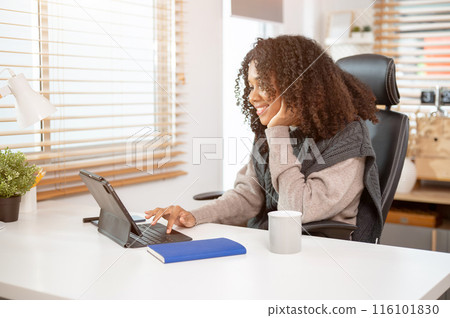 An attractive Black woman is working in her private office, using her digital tablet at her desk. An attractive Black woman is working in her private office, using her digital tablet at her desk. 116101830