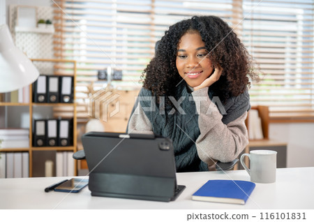 An attractive Black woman is working in her private office, using her digital tablet at her desk. An attractive Black woman is working in her private office, using her digital tablet at her desk. 116101831