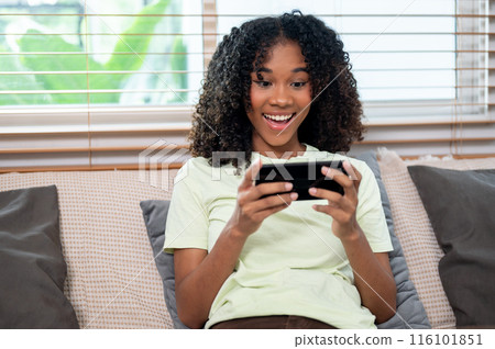 A cheerful, young Black woman is enjoying playing a mobile game on a couch in the living room. 116101851
