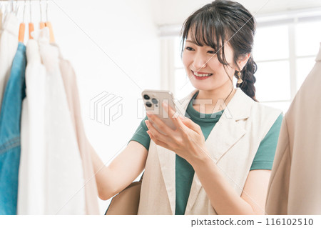 年輕的亞洲女性一邊看著智慧型手機，一邊在服飾店購物 116102510