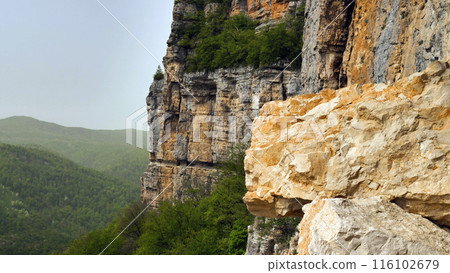 A rugged cliff with contrasting rock layers, with a protrusion of lighter coloured rock in the foreground, a receding cliff edge in the middle ground and a green, forested valley in the background. 116102679