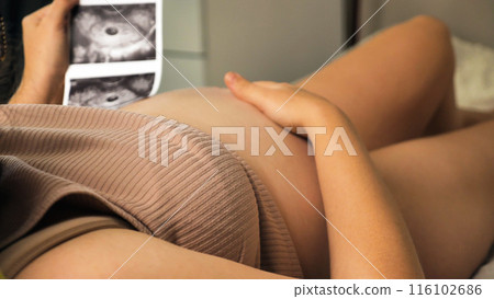 Close-up of a pregnant woman lying in a ribbed brown top, holding an ultrasound image in front of her belly. The mother-to-be is looking at a picture of the baby. Close-up of a pregnant woman lying in a ribbed brown top, holding an ultrasound image in front of her belly. The mother-to-be is looking at a picture of the baby. 116102686