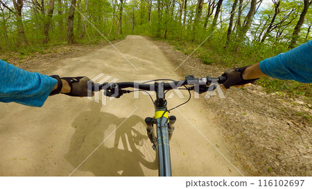 First-person view of a cyclist riding a mountain bike on a dirt trail. The cyclist is wearing a blue shirt and black gloves. The trail is surrounded by lush greenery and trees. First-person view of a cyclist riding a mountain bike on a dirt trail. The cyclist is wearing a blue shirt and black gloves. The trail is surrounded by lush greenery and trees. 116102697