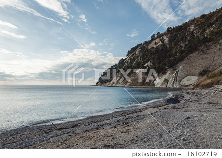 A serene coastal scene with a rocky beach, clear blue water, and a steep cliffside covered in trees. A few people are scattered along the shore, and a tent is set up near the water. 116102719