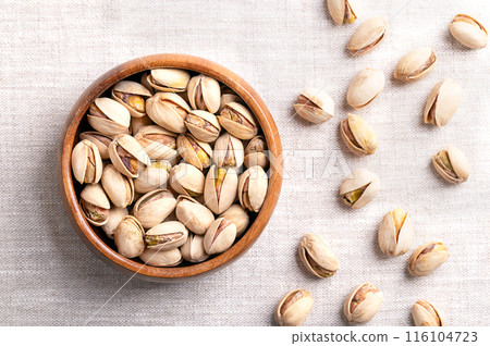 Pistachios in their shells, roasted and salted, in a wooden bowl on linen fabric. Crunchy roasted fruits of Pistacia vera, with opened shells and seeds within. Ready-to-eat snack food. Close-up. Photo Pistachios in their shells, roasted and salted, in a wooden bowl on linen fabric. Crunchy roasted fruits of Pistacia vera, with opened shells and seeds within. Ready-to-eat snack food. Close-up. Photo 116104723
