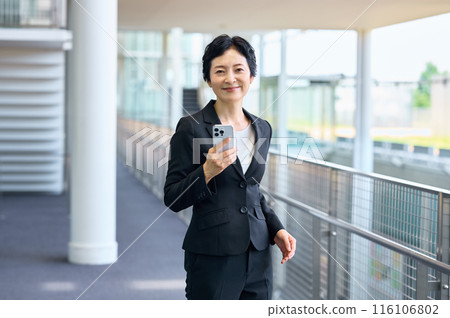 A businesswoman in a suit standing with a smartphone in an office corridor A businesswoman in a suit standing with a smartphone in an office corridor 116106802