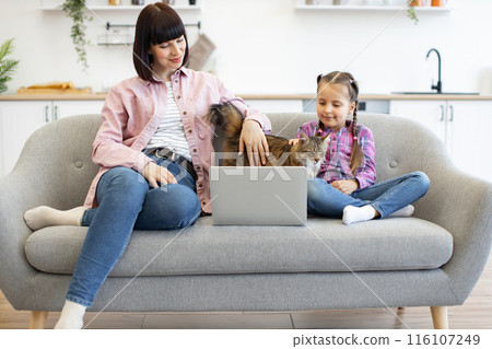 Mother and daughter with laptop and cat on sofa at home 116107249