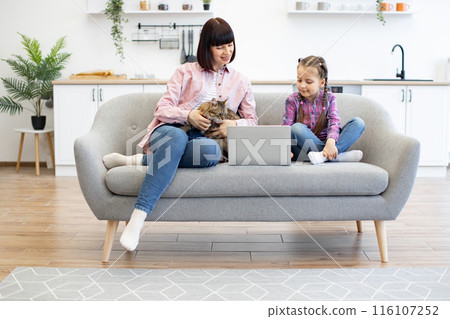 Mother and daughter sitting on sofa with cat using laptop 116107252