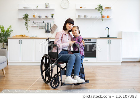 Mother in wheelchair hugging daughter in bright kitchen Mother in wheelchair hugging daughter in bright kitchen 116107388