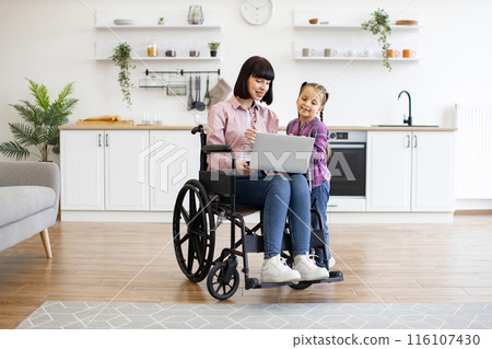 Mother in wheelchair using laptop with daughter in kitchen Mother in wheelchair using laptop with daughter in kitchen 116107430