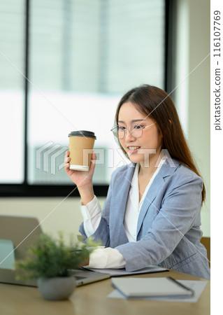 Young businesswoman in eyeglasses using laptop and having coffee at desk in a modern office 116107769