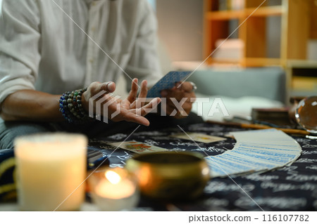 Close up shot of a fortune teller wearing several beaded bracelets holding a tarot card near various spiritual items Close up shot of a fortune teller wearing several beaded bracelets holding a tarot card near various spiritual items 116107782