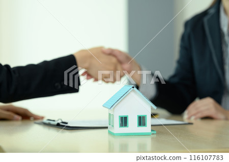 Small house model on wooden table with real estate agent shaking hands with his customer in background Small house model on wooden table with real estate agent shaking hands with his customer in background 116107783