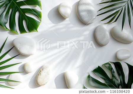 Smooth white stones rest on a pristine white background, flanked by lush green monstera and palm leaves, the serene composition evokes a sense of tranquility and natural beauty, spa concept 116108420