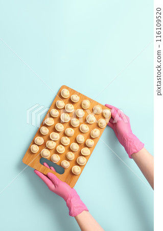 Chef's hands in rubber gloves hold a wooden board with neatly arranged dumplings on a blue background, flat lay and copy space. Chef's hands in rubber gloves hold a wooden board with neatly arranged dumplings on a blue background, flat lay and copy space. 116109520