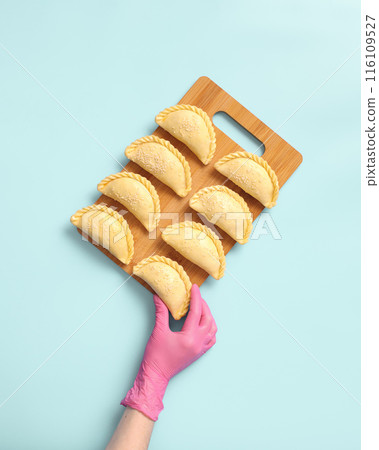 A baker wearing a rubber glove arranges raw samosas on the table. These are frozen ready-to-cook traditional Central Asian delicacies. 116109527
