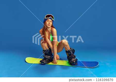 Girl combining summer swimwear and winter sports gear, ready for a unique snowboarding experience against blue studio background Girl combining summer swimwear and winter sports gear, ready for a unique snowboarding experience against blue studio background 116109870