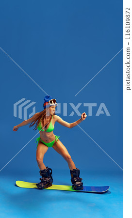 Stylish woman in bikini and winter sports gear, demonstrating her adventurous spirit and love for snowboarding against blue studio background 116109872