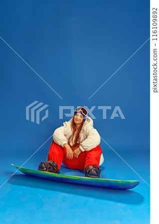 Woman combining summer and winter attire, relaxing with her snowboard, sitting against blue studio background. Extreme sports ads 116109892