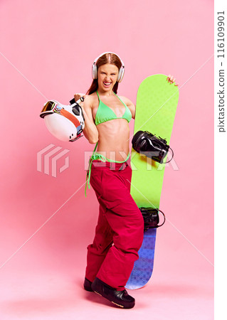 Happy and emotional young girl with tanned body, posing in winter clothes and bikini top, with snowboarding equipment against pink studio background Happy and emotional young girl with tanned body, posing in winter clothes and bikini top, with snowboarding equipment against pink studio background 116109901