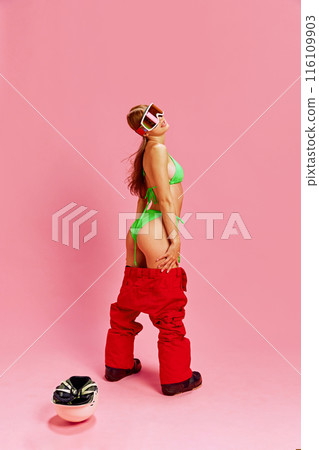 Young woman in a green bikini and red snow pants, embracing a playful and confident pose against pink studio background. Holiday fun Young woman in a green bikini and red snow pants, embracing a playful and confident pose against pink studio background. Holiday fun 116109903