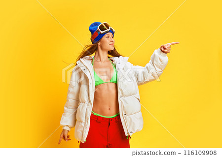 Happy woman in unique mix of winter sports attire and summer wear, choosing her travelling destination against bright yellow studio background Happy woman in unique mix of winter sports attire and summer wear, choosing her travelling destination against bright yellow studio background 116109908